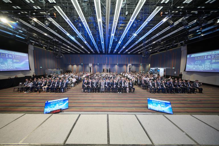 A wide-angle shot of a large conference hall filled with attendees seated in rows, facing a stage with a massive screen displaying the "E&M I&T Day 2026" event branding.